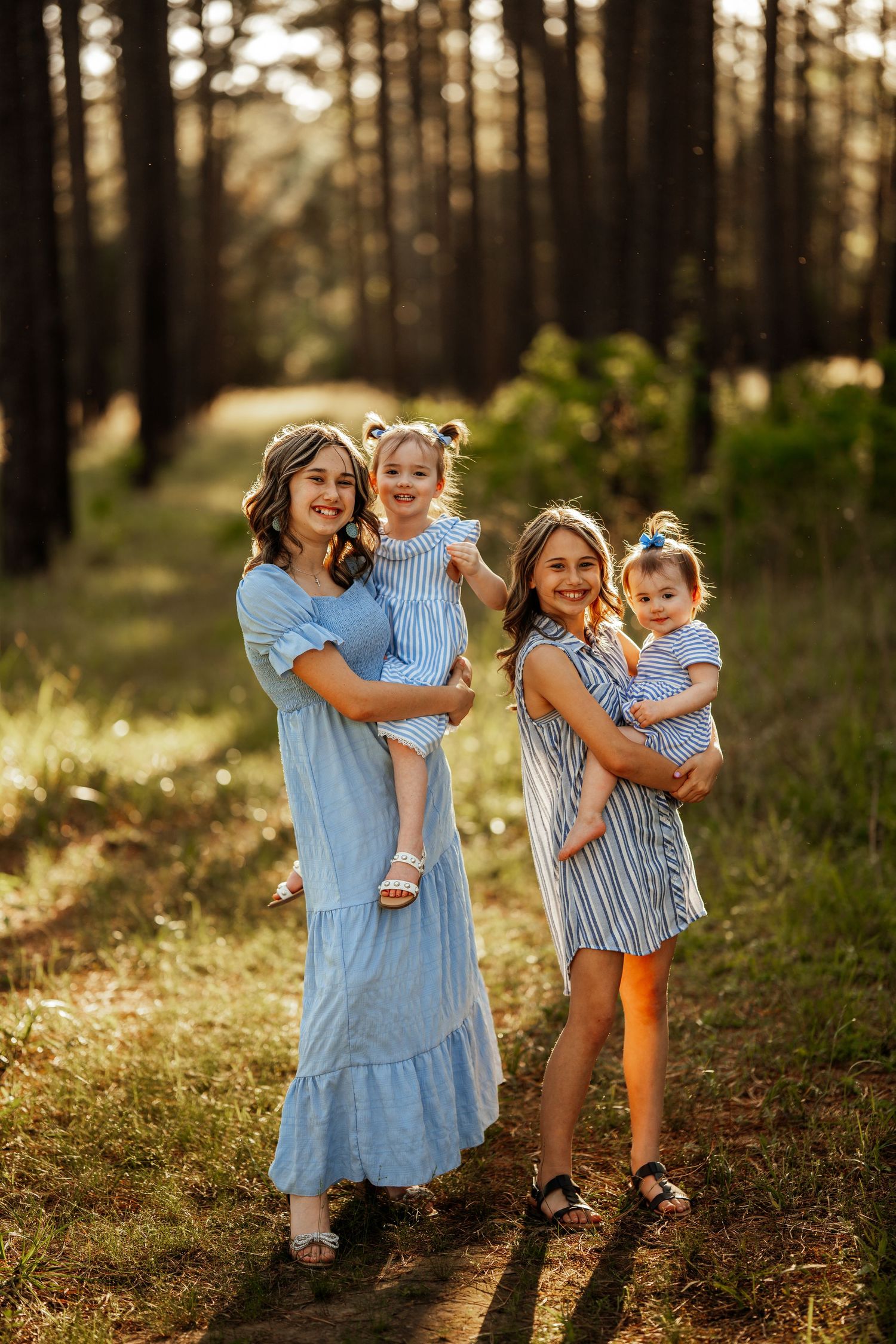 Four people in coordinating blue and striped dresses pose together on a sunlit forest path at golden hour.