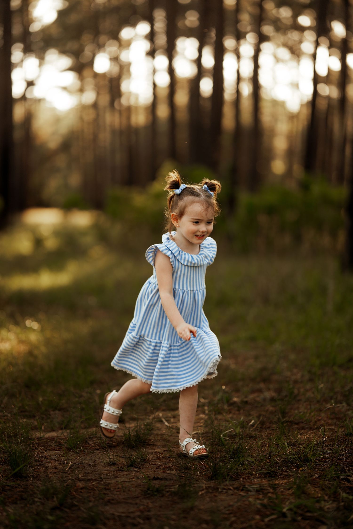 Young child in blue striped dress dances in a sunlit forest during golden hour.