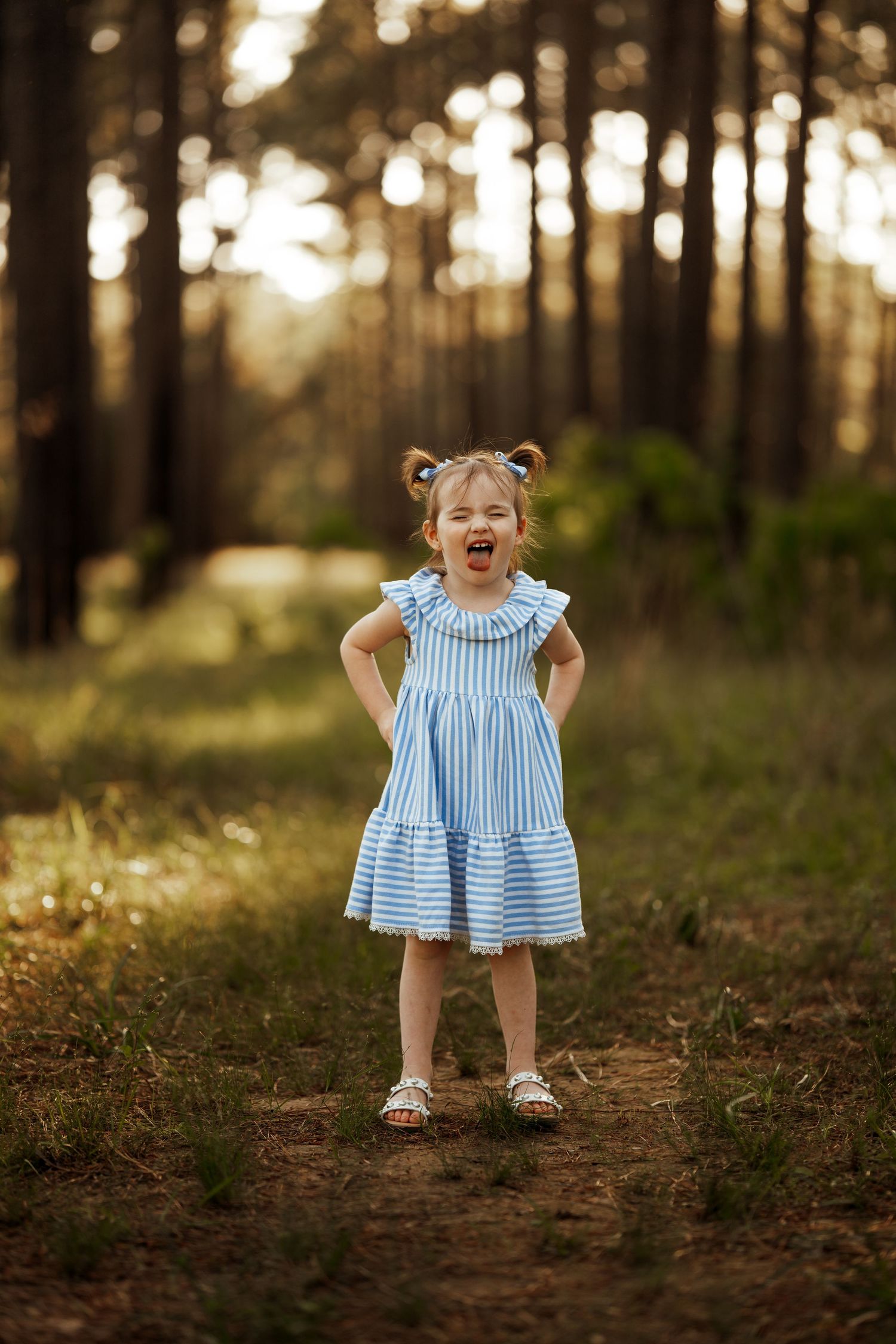 A child wearing a blue and white striped dress stands on a dirt path in a sunlit forest at golden hour.