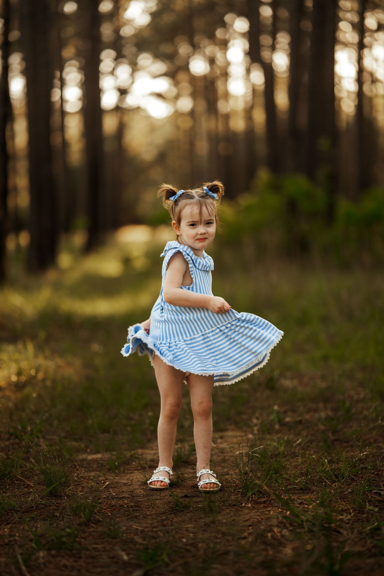 A small child in a blue striped dress twirls on a forest path during golden hour.