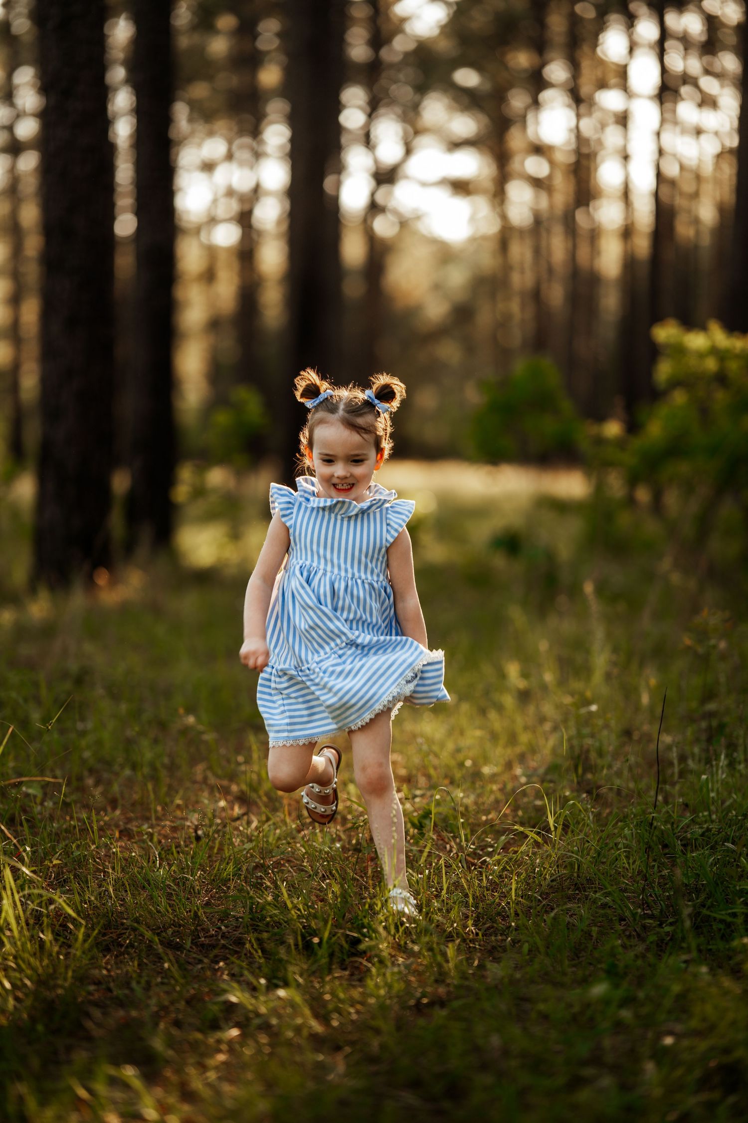 A child wearing a blue striped dress runs playfully through a sunlit forest at golden hour.