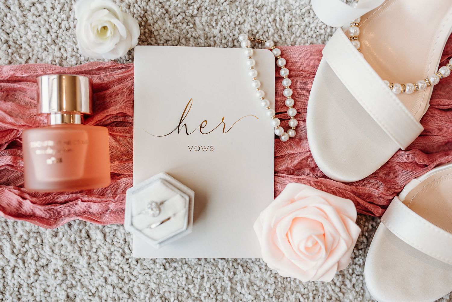 Wedding details flatlay with perfume, rings, and pink roses on textured background.