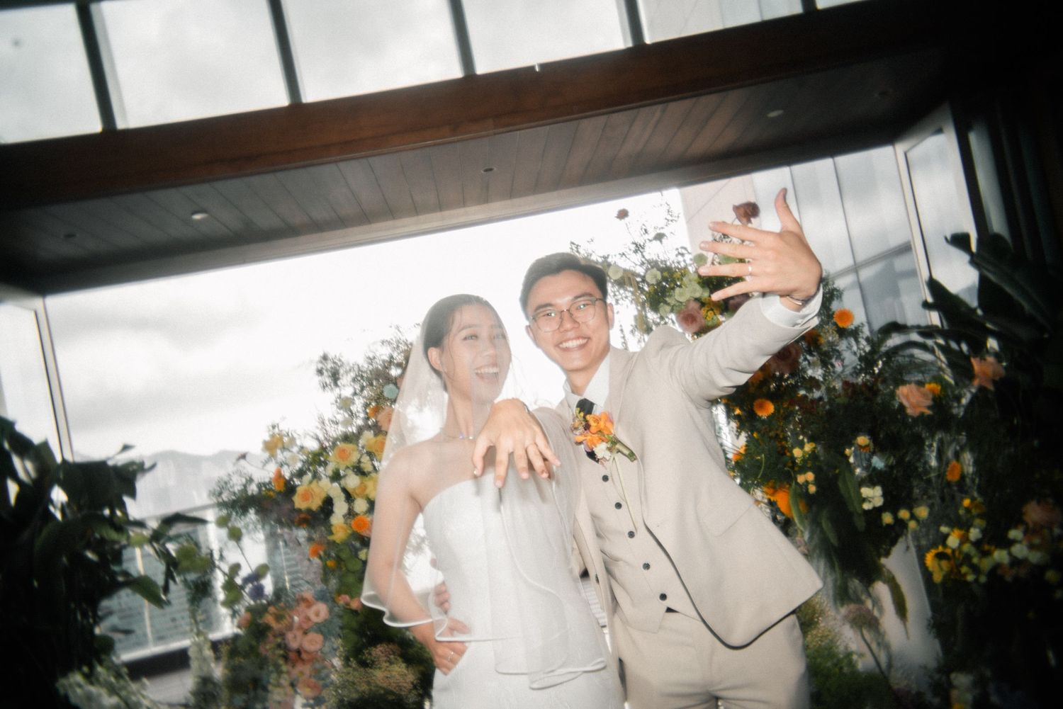 A couple in white and beige wedding attire celebrate with raised arms under floral decorations.