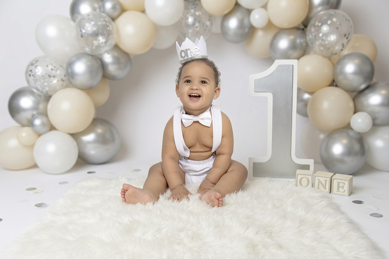 First birthday setup with silver and cream balloons and baby in white chair.