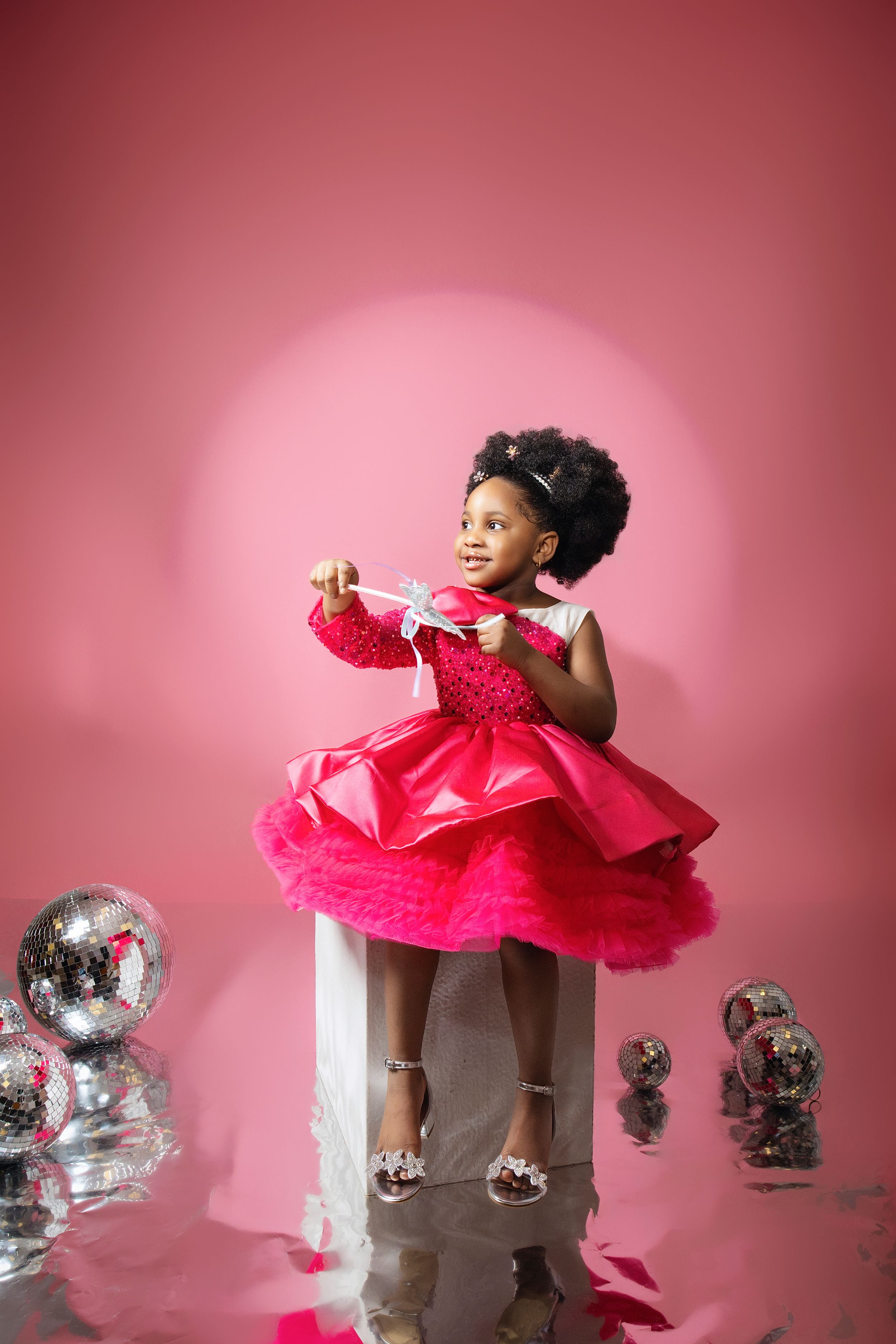 Young child in bright pink tulle dress playing with silver decorations against pink backdrop.