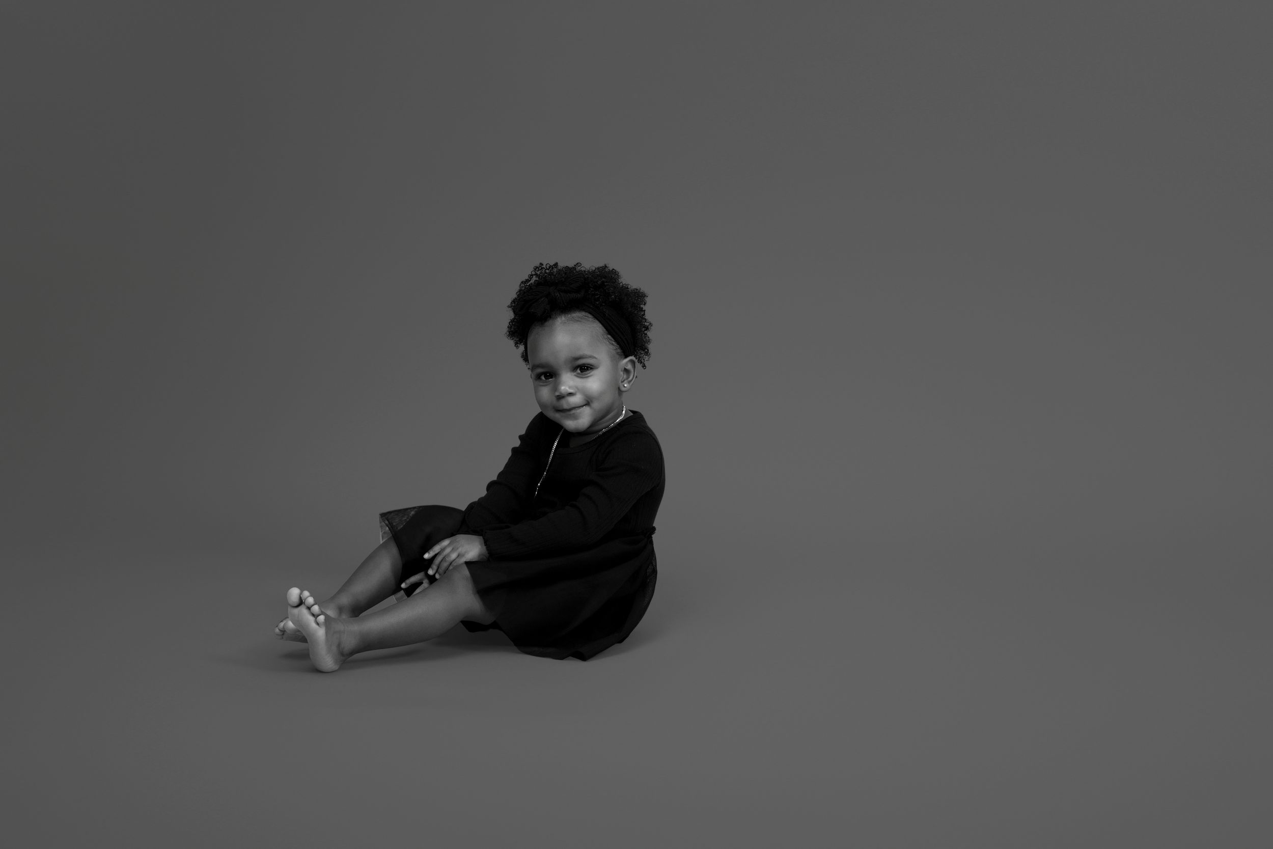 Artistic black and white portrait of child sitting on floor in dark outfit.