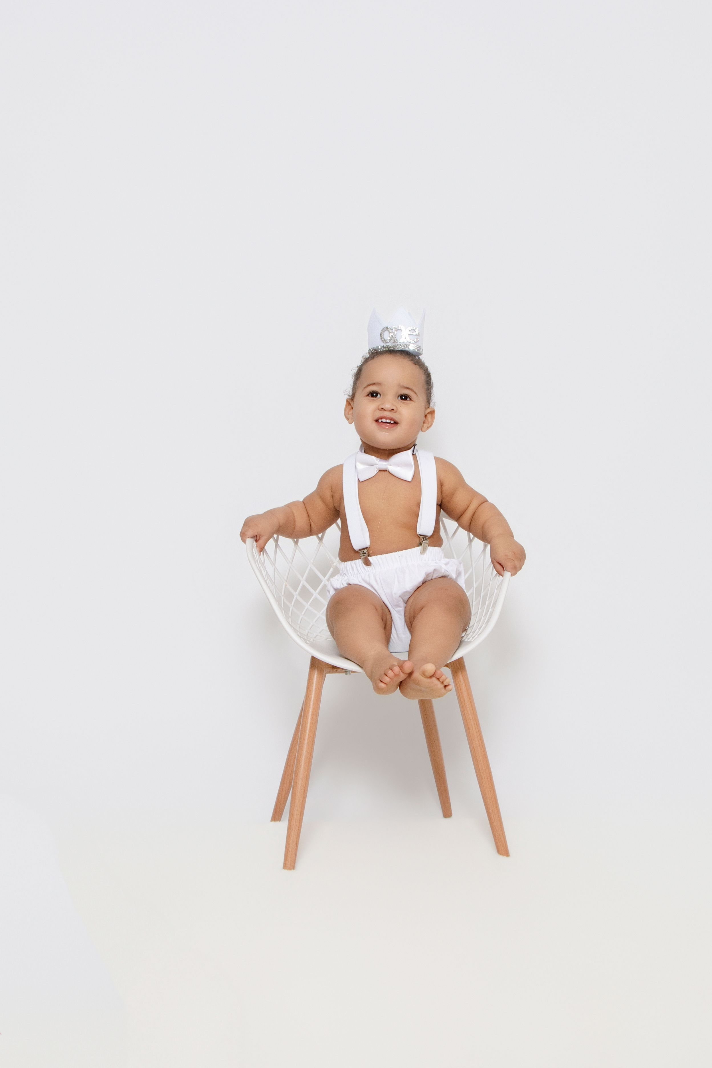 Baby wearing white outfit sits in modern wooden high chair against white backdrop.