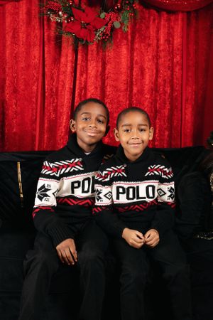 Two children wearing matching Polo sweaters sitting on couch with red backdrop and holiday decor.