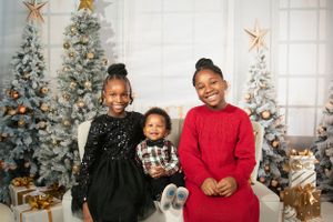 Three children posing on couch with Christmas trees, decorations and stuffed animal in holiday setting.