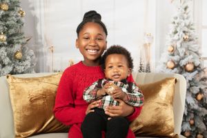 Two children hugging and smiling on couch with Christmas trees and decorations behind them.