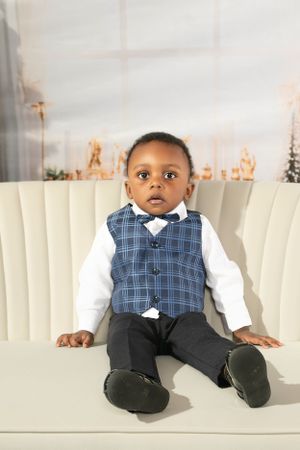 Young child in plaid vest and bowtie sitting on white couch with Christmas decor in background.