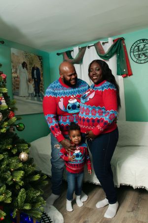 Family wearing matching red and blue Christmas sweaters poses by decorated tree in mint green room.