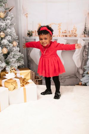 Young child in red dress and bow twirls by Christmas tree and wrapped presents.