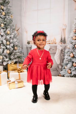 Young child in red dress standing by Christmas trees and presents in festive holiday setting.