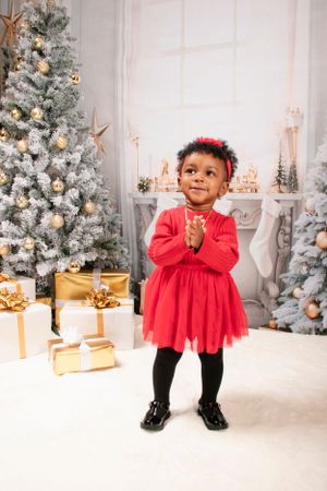 Child in red dress smiling and clapping hands in front of decorated Christmas trees and gifts.