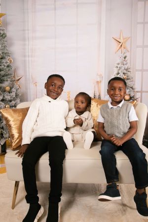 Three children sitting on couch in front of Christmas trees in festive holiday photo setting.
