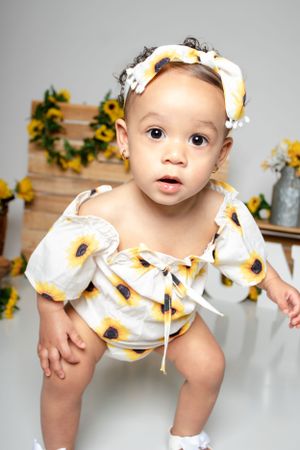 Baby in sunflower print outfit against grey background with floral decorations.