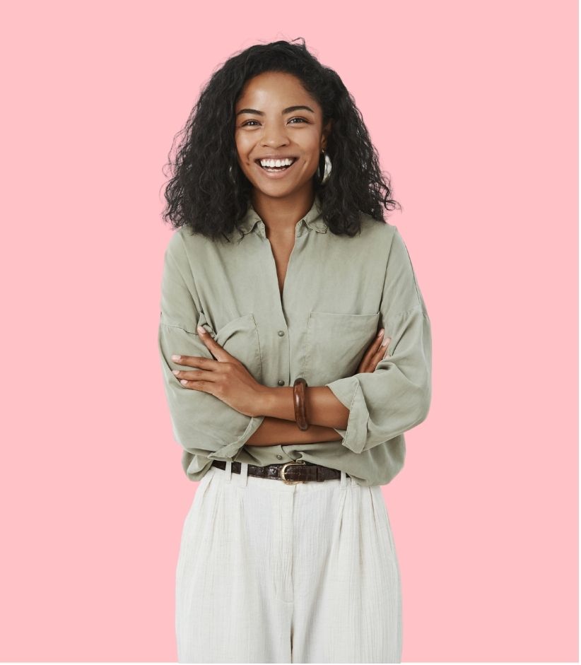 Professional portrait of a person with curly hair wearing a sage green blouse and white pants against a coral background.