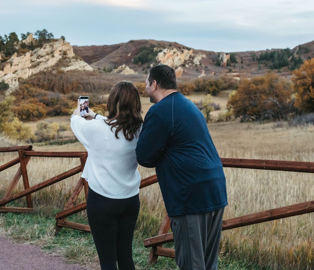 Two people stand at a rustic railing overlooking scenic mountain views during autumn.