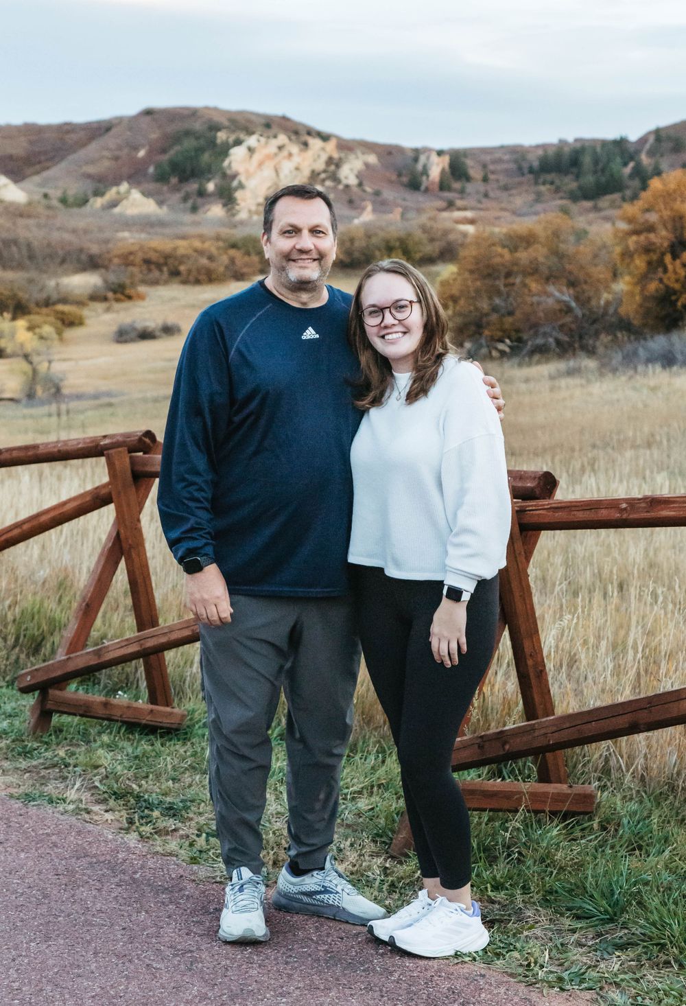A pair stands together smiling on a hiking trail with rustic wooden fencing and mountain scenery in the background.
