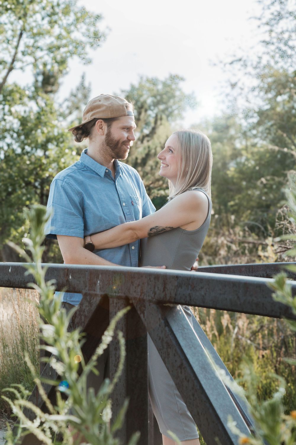 A couple shares a romantic moment together on a wooden bridge surrounded by sunlit greenery and tall grass.