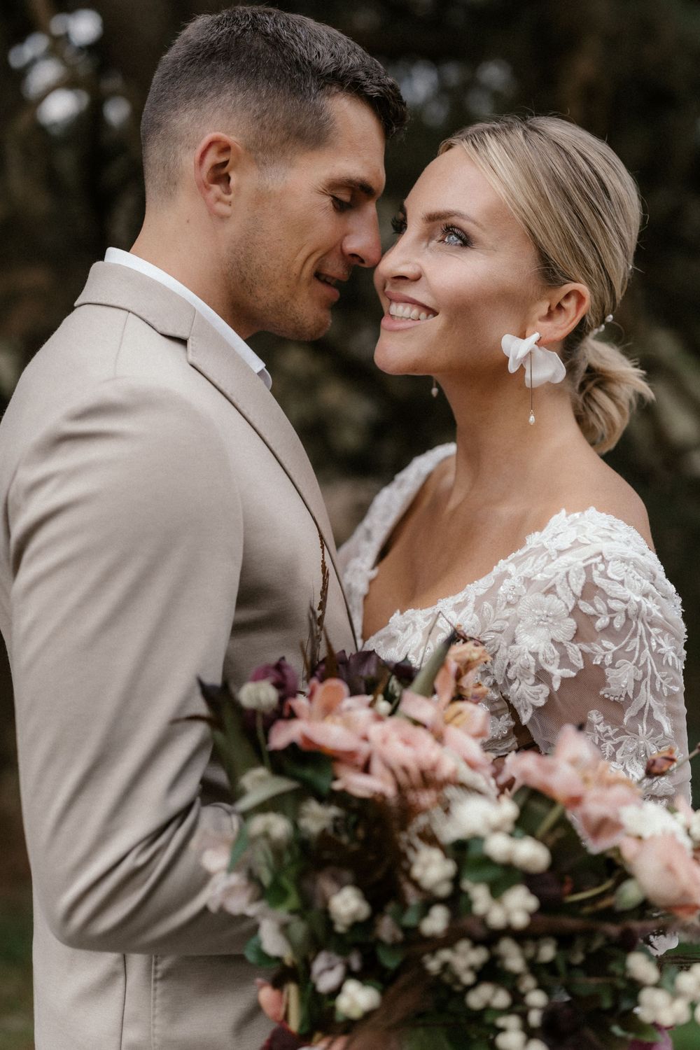 Frisch vermähltes Paar in beigem Anzug und Spitzenkleid teilt romantischen Moment mit rosa Blumen.