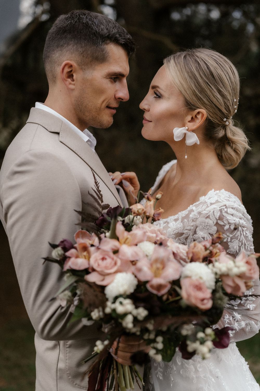 Braut im Spitzenkleid und Bräutigam im hellbraunen Anzug teilen intime Momente mit einem rosa Blumenstrauß.