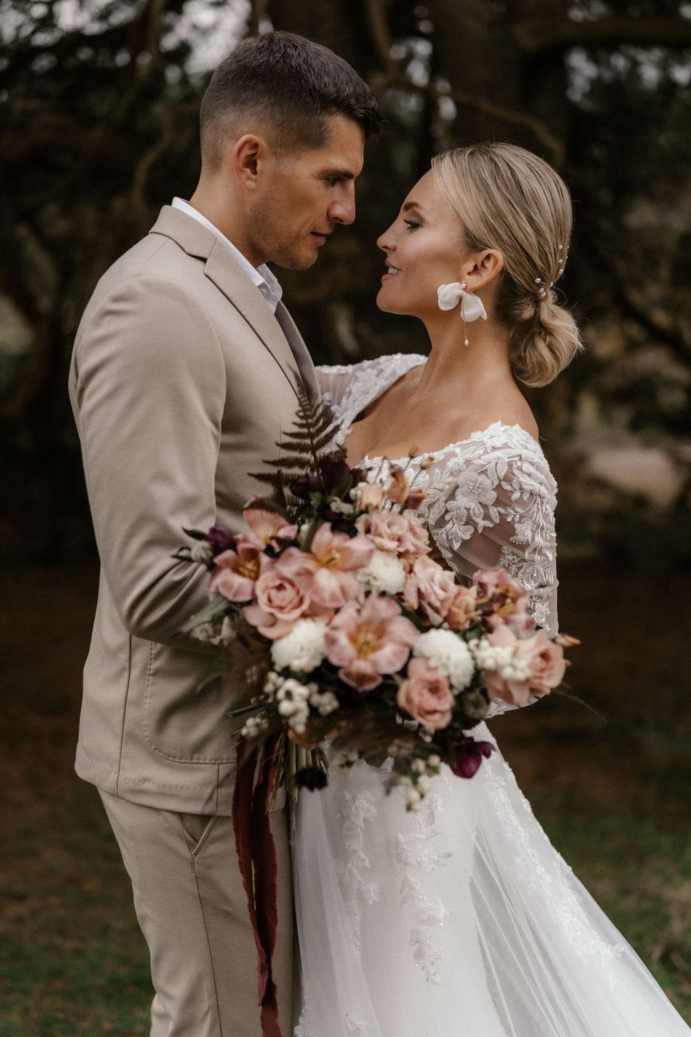 Elegantes Paar in beigem Anzug und Spitzenkleid posiert mit rosa Rosenstrauß während einer Hochzeit im Freien.