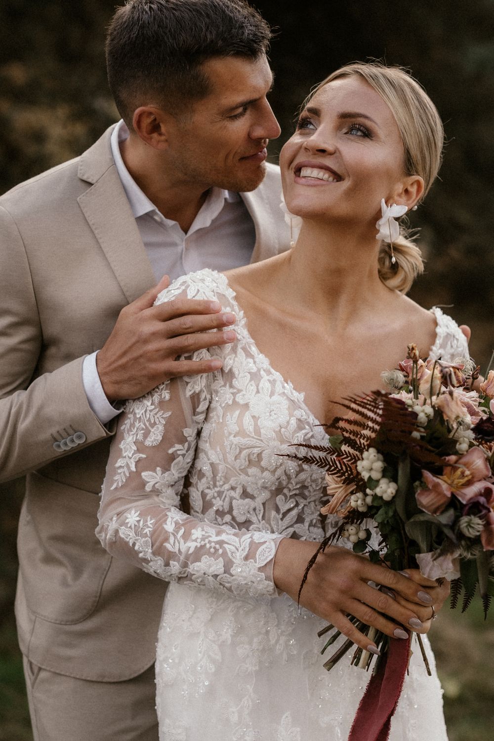 Romantisches Hochzeitspaar umarmt sich freudig und hält dabei einen wunderschönen rosa-weißen Blumenstrauß in den Händen.