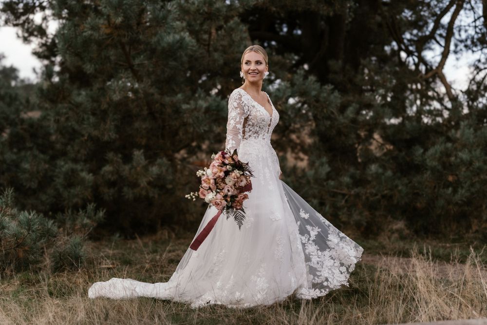 Braut im Spitzenbrautkleid wirbelt im Freien herum und hält einen burgunder- und rötlichen Blumenstrauß in der Hand.