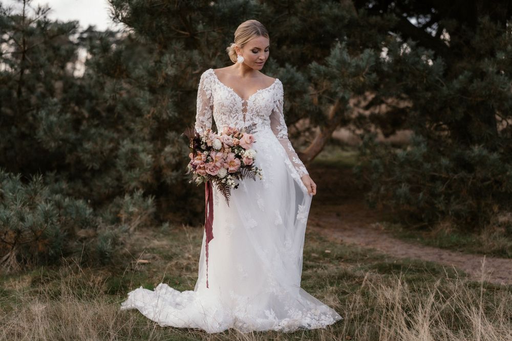 Solo-Brautporträt mit Spitzenkleid und burgunderfarbenem Blumenstrauß im Freien.