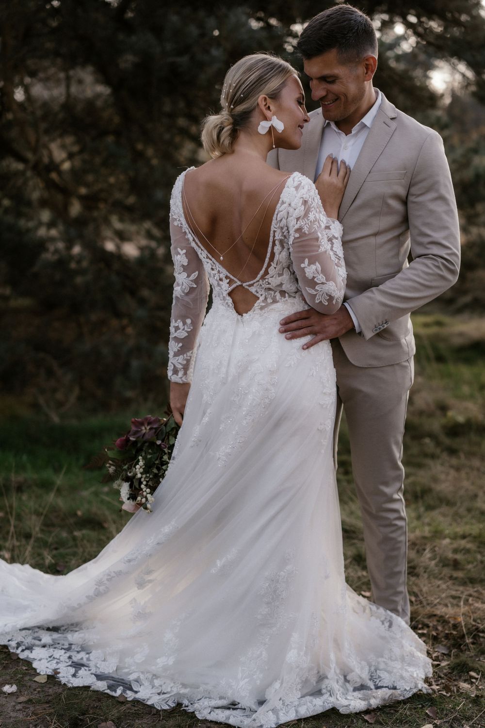 Romantisches Hochzeitsporträt im Freien mit hellem Spitzenkleid und hellbraunem Anzug im warmen Licht des Sonnenuntergangs.
