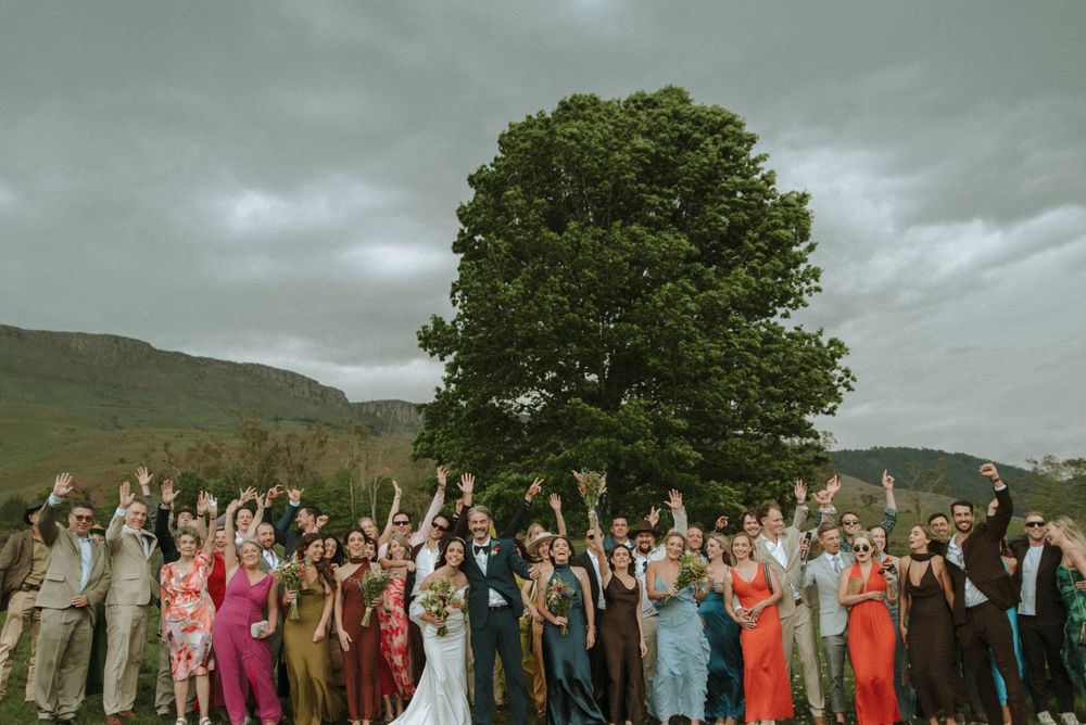 A wedding party poses outdoors in front of a dramatic cloudy sky and mountainous landscape.