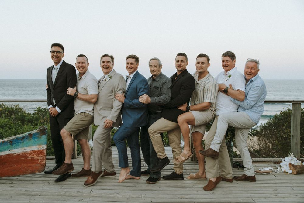 Group of nine men posing and laughing on a seaside deck at sunset, dressed in smart casual attire.