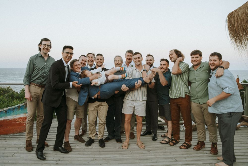 Group of men smiling and lifting one person at an outdoor beach celebration party.