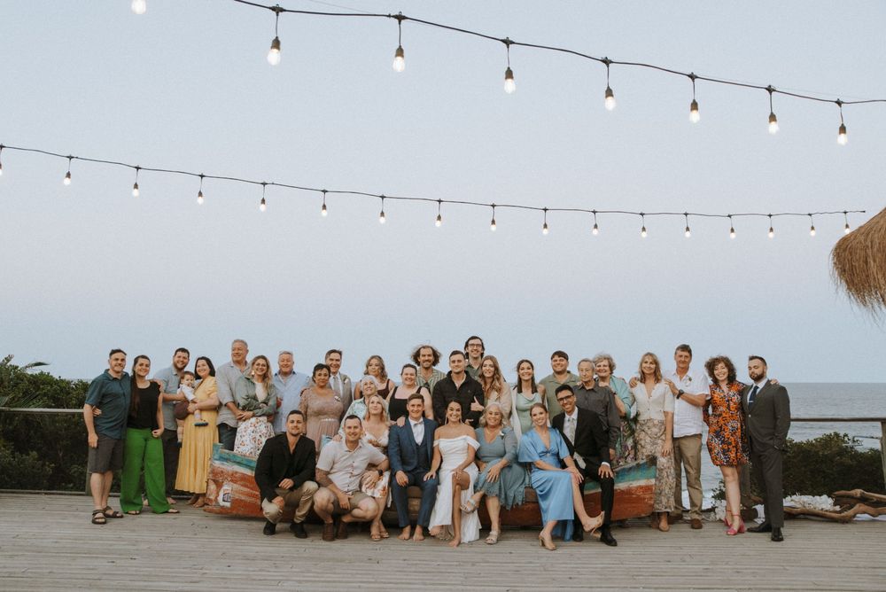 Large wedding group photo outdoors at dusk with string lights overhead near ocean backdrop.
