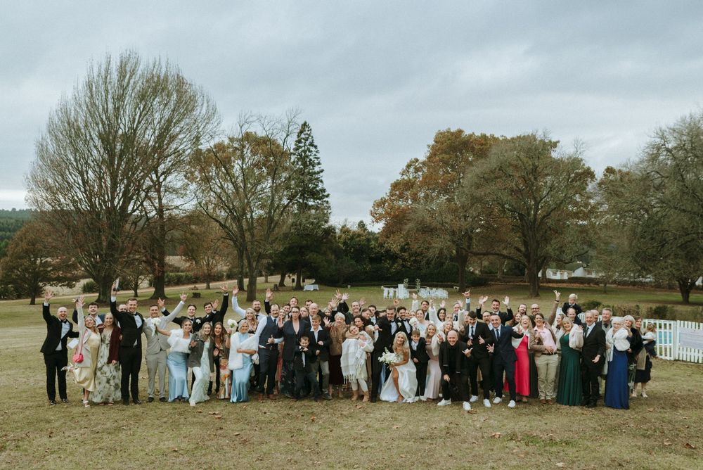 Large wedding group photo outdoors on green lawn with bare trees, guests cheering and raising arms in celebration.