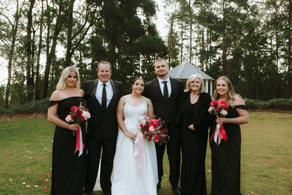 Wedding party in formal attire poses outdoors on grassy field with trees in background.