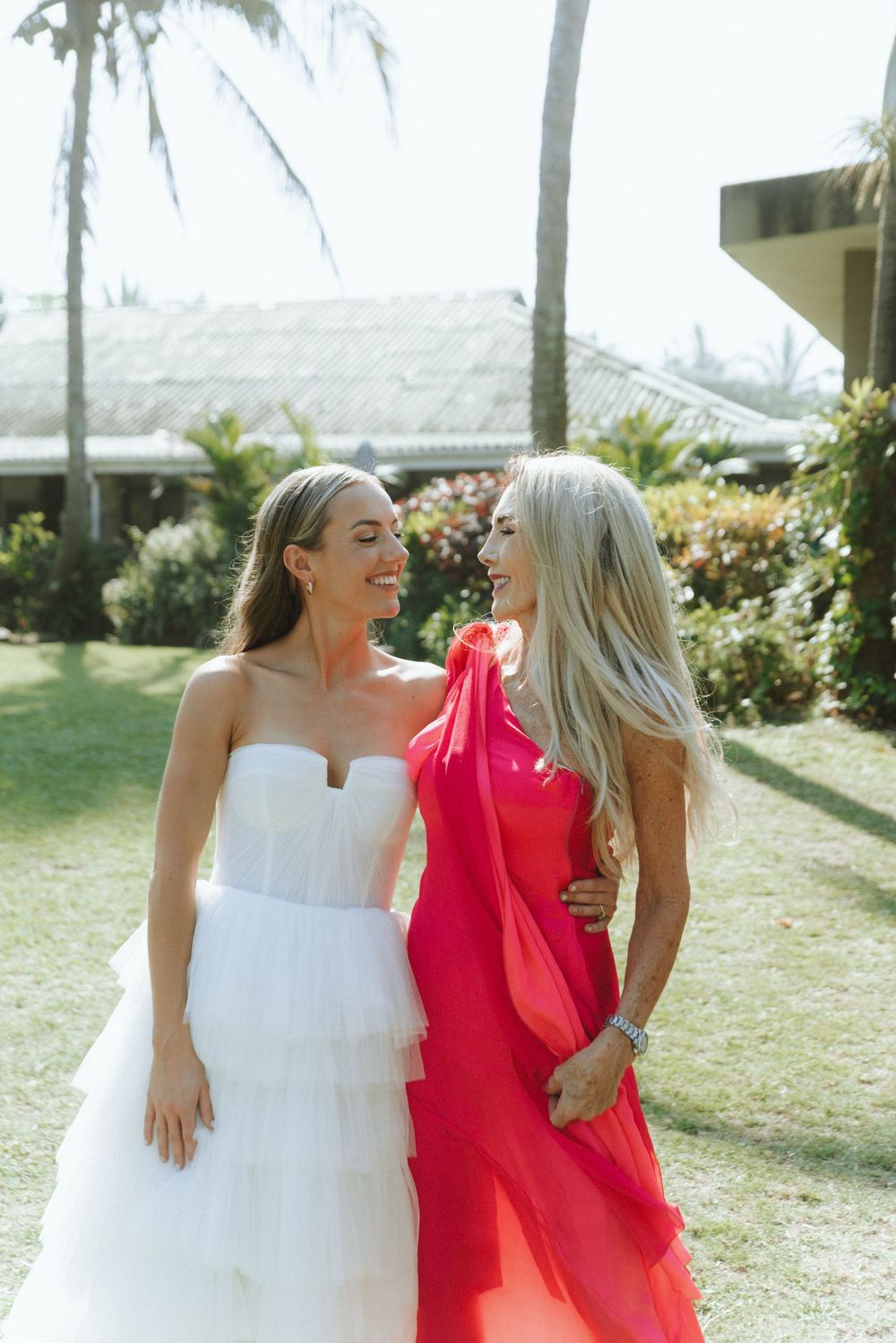 Two friends in white and coral dresses share laughter while walking together outdoors on a sunny day.