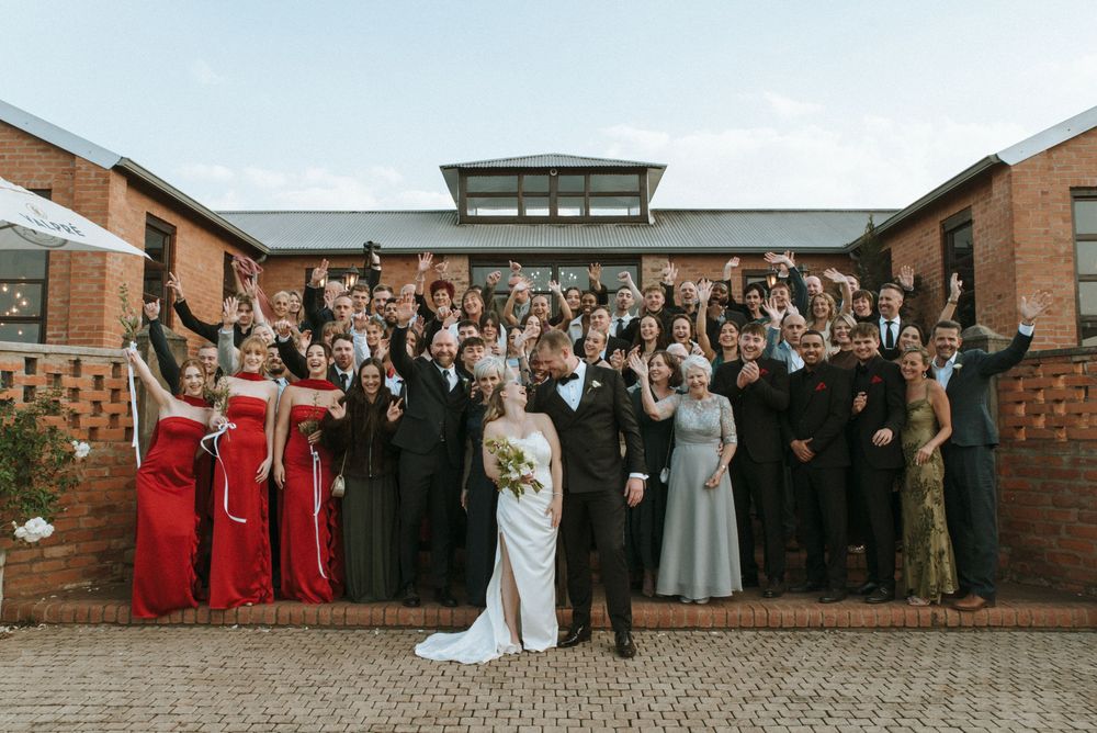 A bride and groom pose with their large wedding party and guests outside a brick venue, all celebrating joyfully.