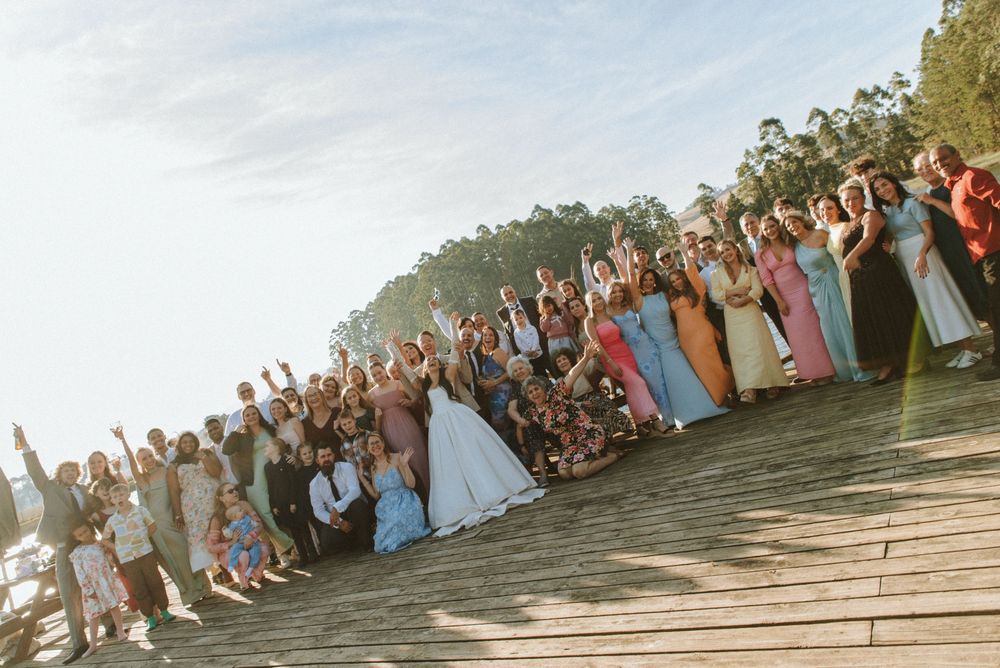 Large wedding group photo with bride in white gown, guests in colorful attire, waving on wooden steps outdoors.