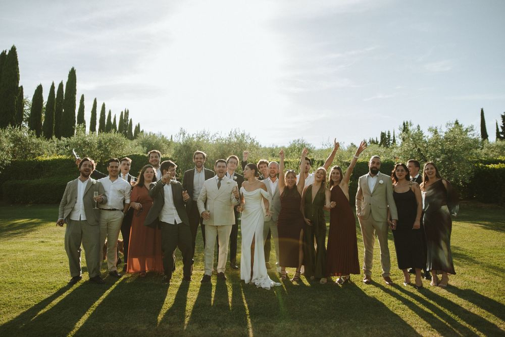 Wedding party celebrating outdoors in Tuscany, bride and groom surrounded by guests in elegant attire on lush green lawn.