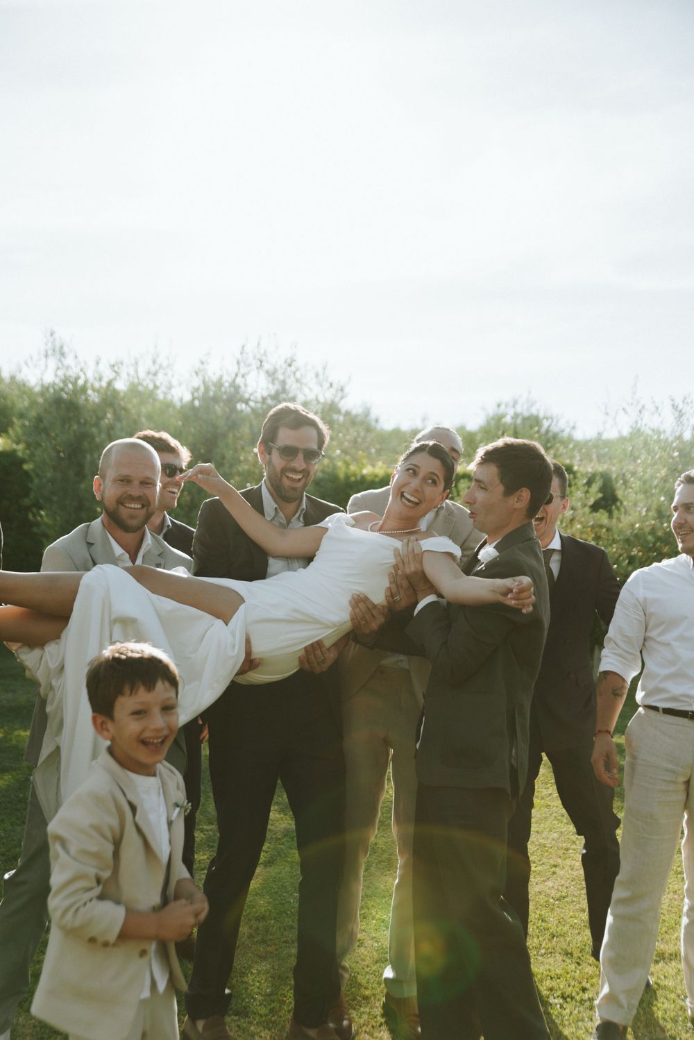 Groomsmen joyfully lifting smiling bride outdoors at wedding celebration with young ring bearer and lush greenery in background.