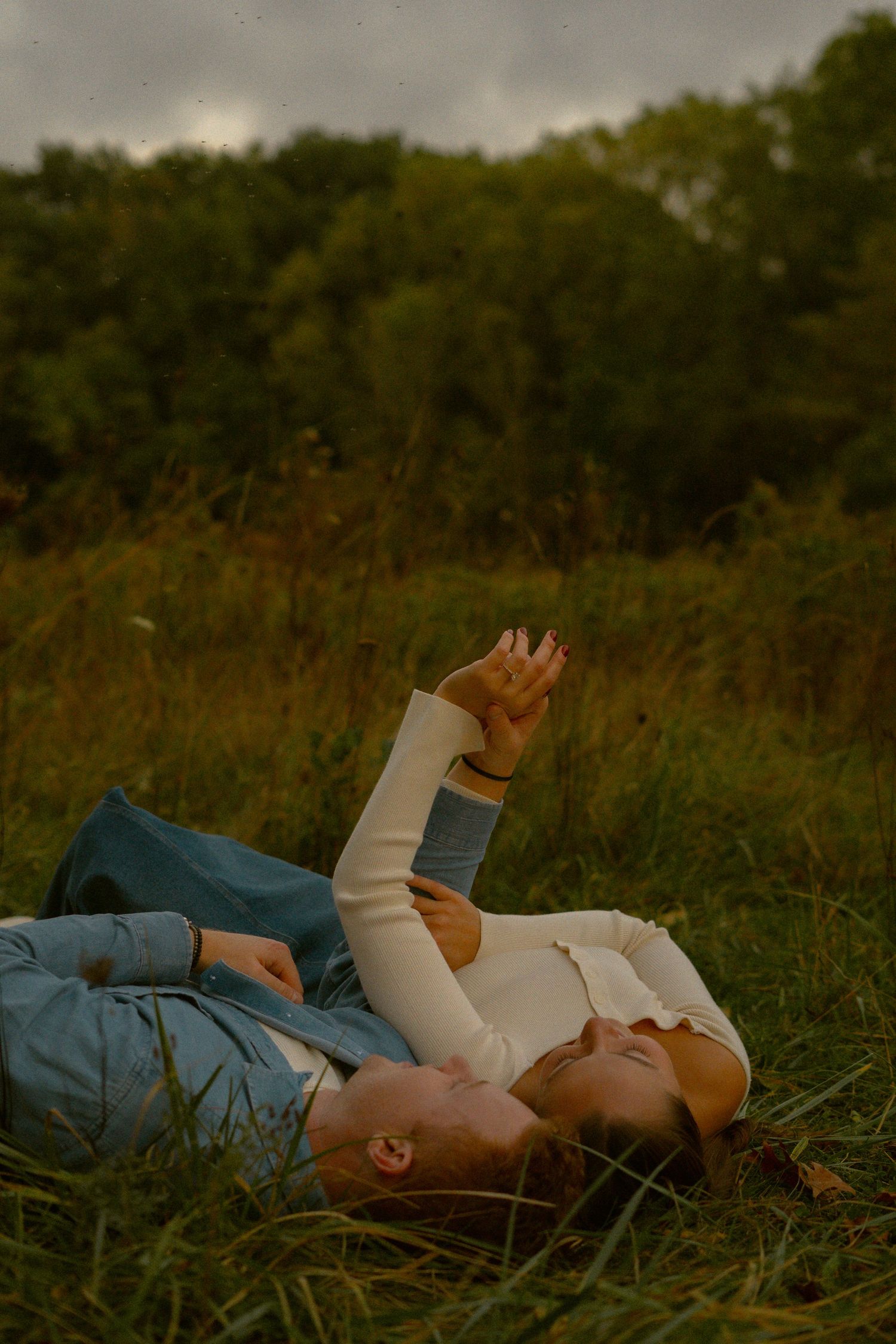 Couple lying in tall grass at dusk reaching towards the sky in a peaceful moment.