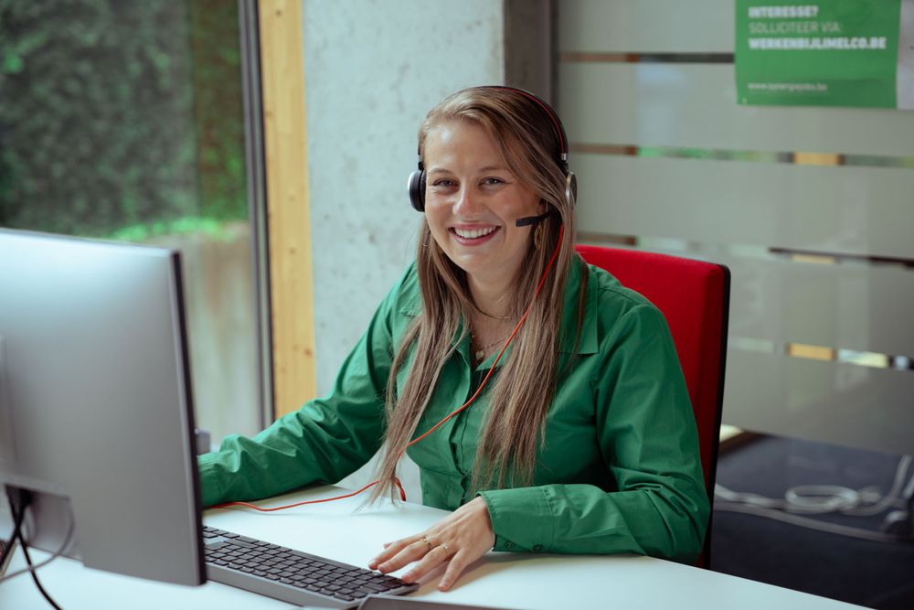 Woman laughing while wearing headphones, working at a computer in a modern office environment in Hasselt.
