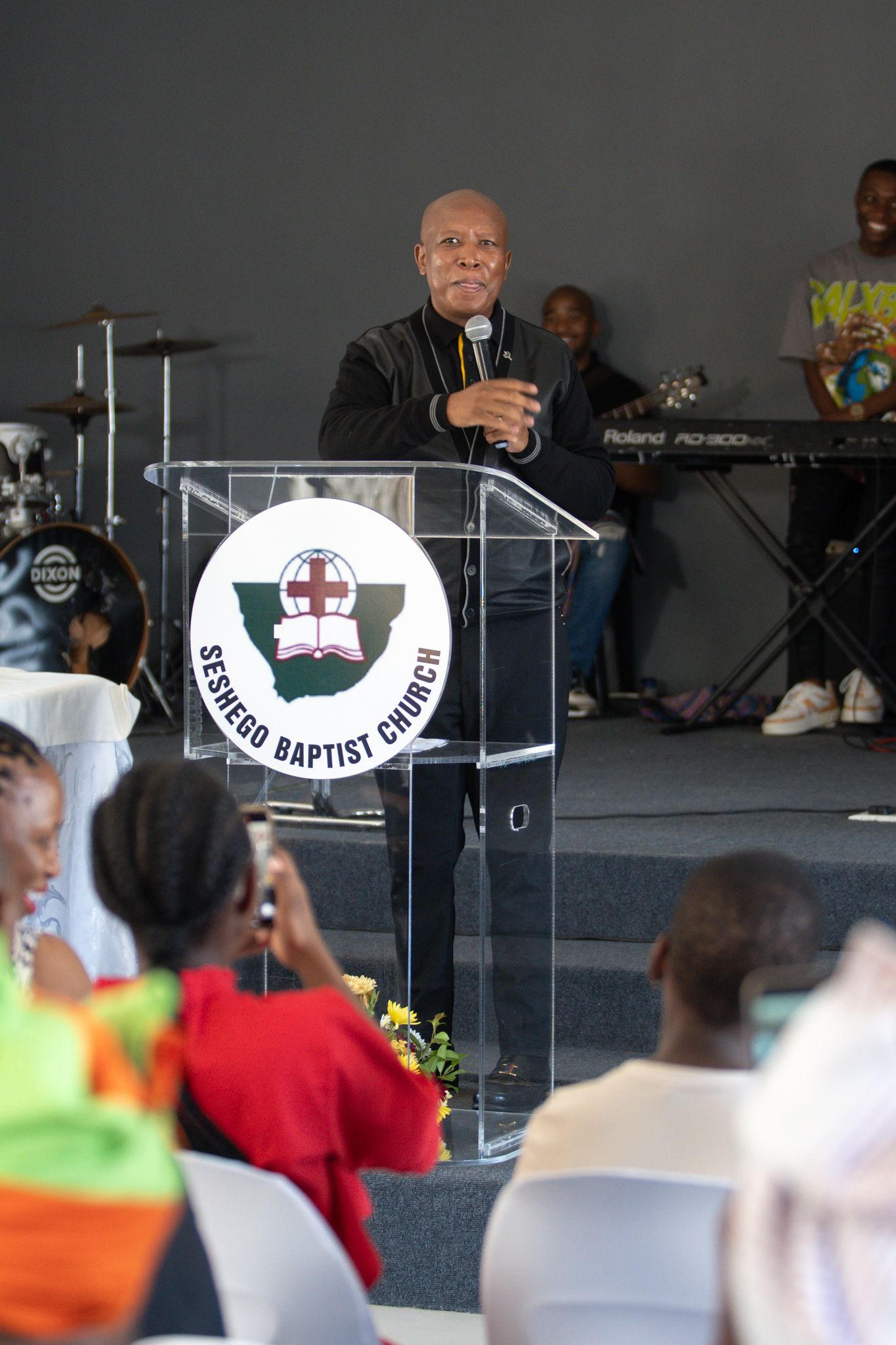 Speaker at podium delivers message during Baptist church service with musicians visible in background.