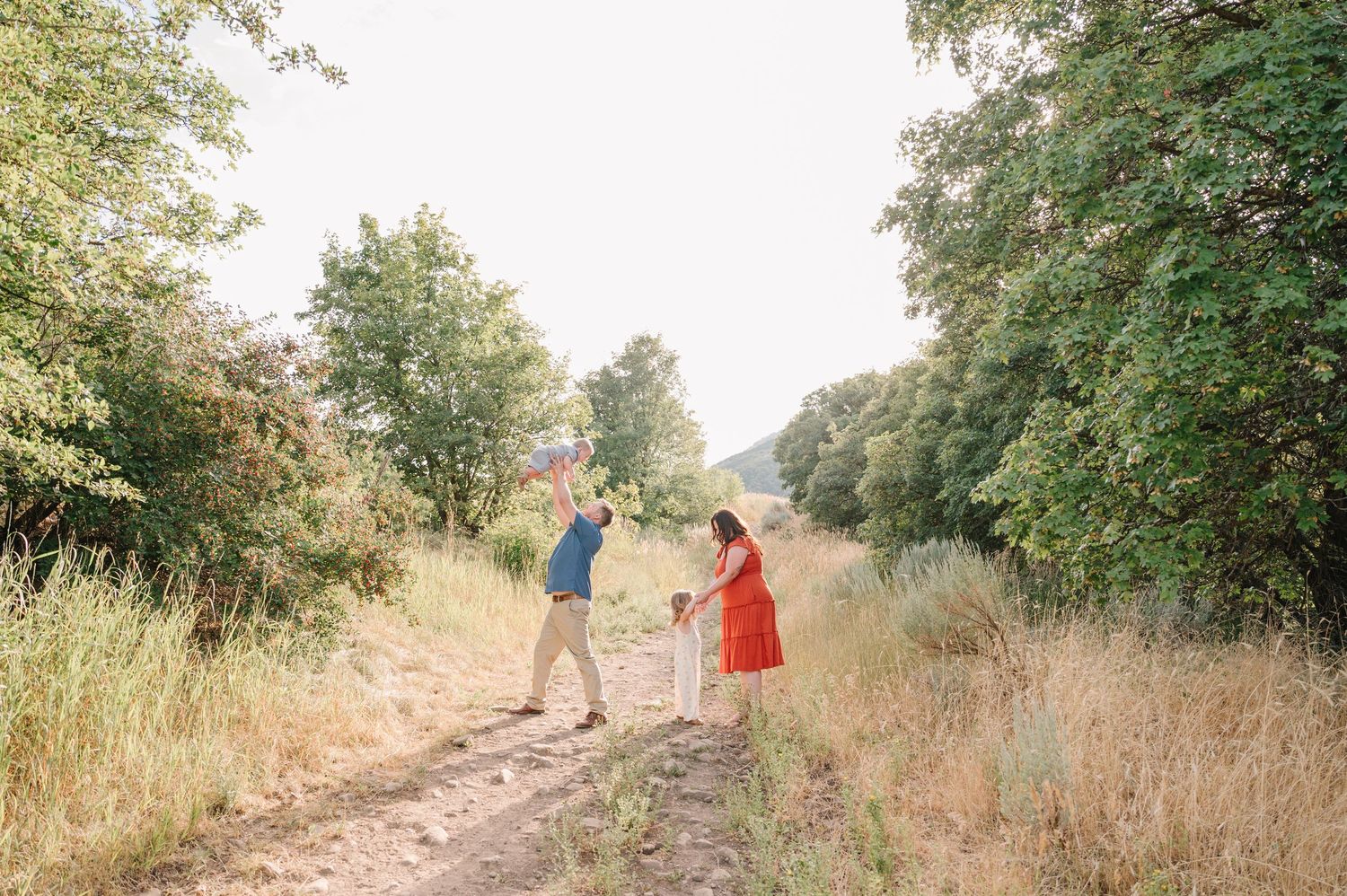 Two people in a serene outdoor setting walk along a dirt trail surrounded by golden grass and green trees on a sunny day.