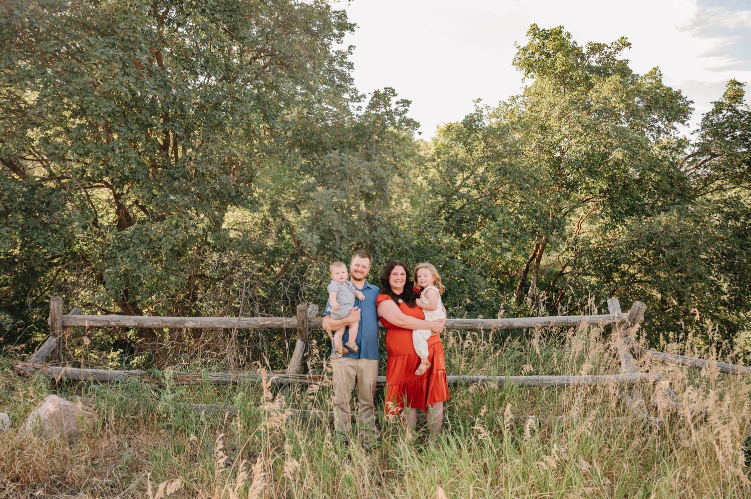A group standing together in a red dress and blue shirt against a rustic wooden fence in a meadow at sunset.