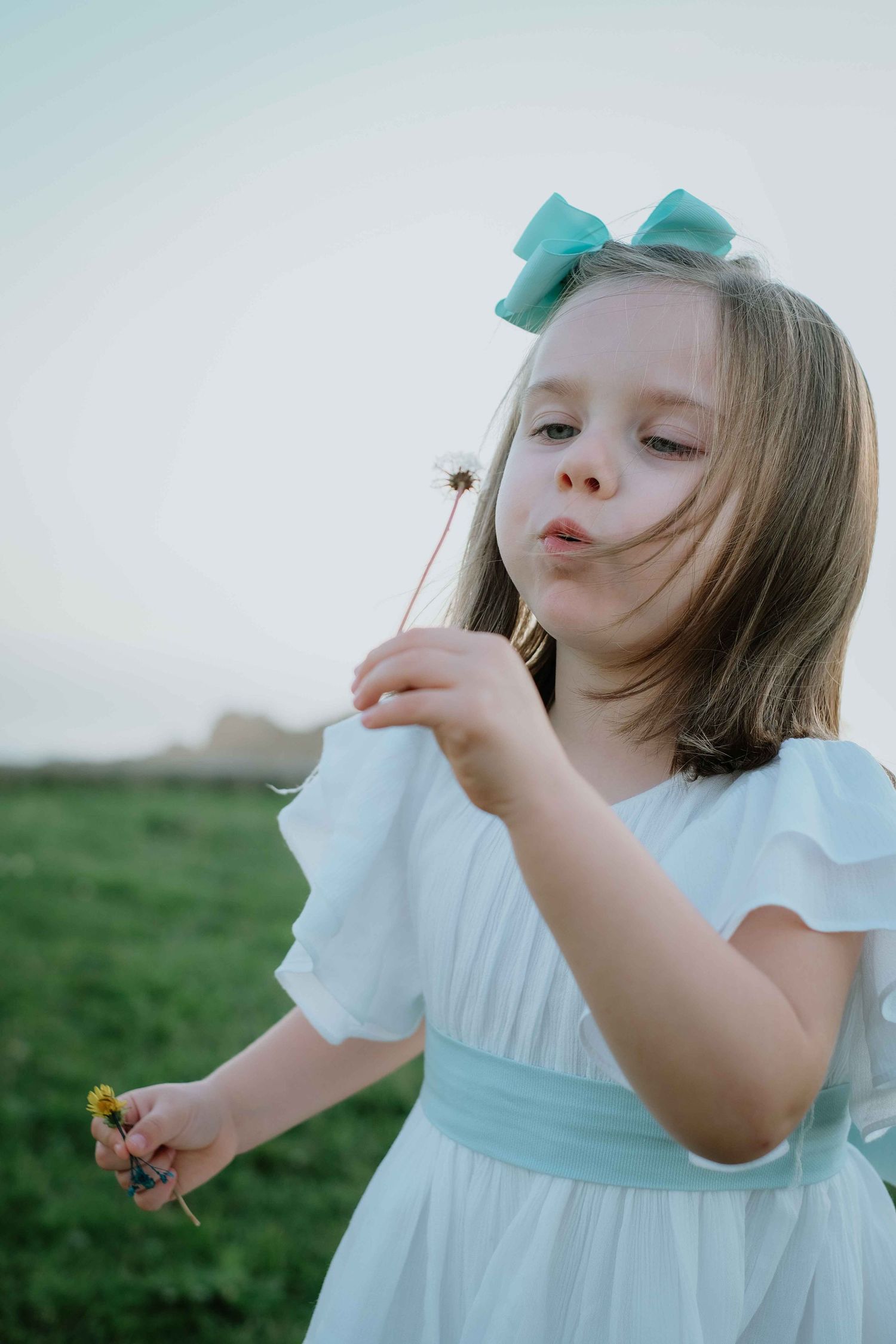 Child in white dress with blue bow playing outdoors in soft lighting.