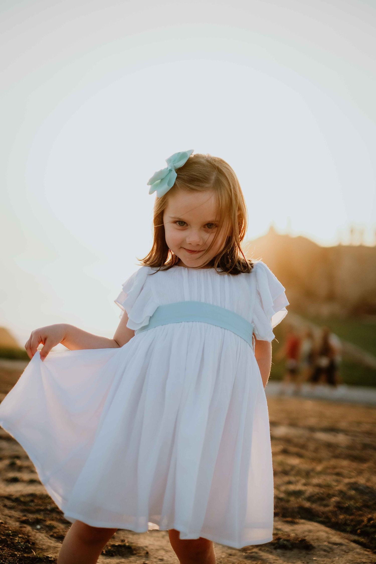 Child twirling in white dress with blue bow against sunset backdrop.
